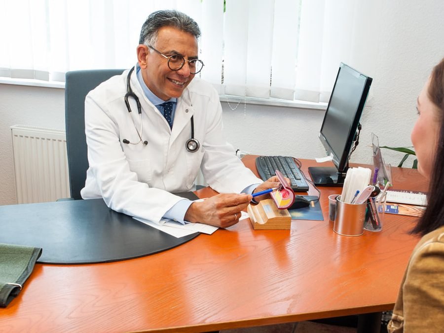 Arzt im weißen Kittel im Gespräch mit einer Patientin am Schreibtisch, auf dem Tisch ein Blutdruckmessgerät